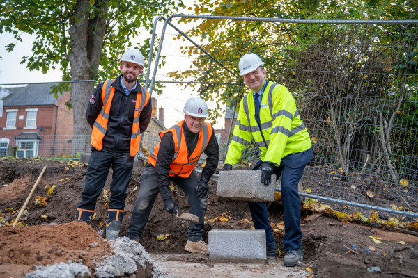 Three people wearing high visibility jackets, hard hats and gloves place a brick on a building site